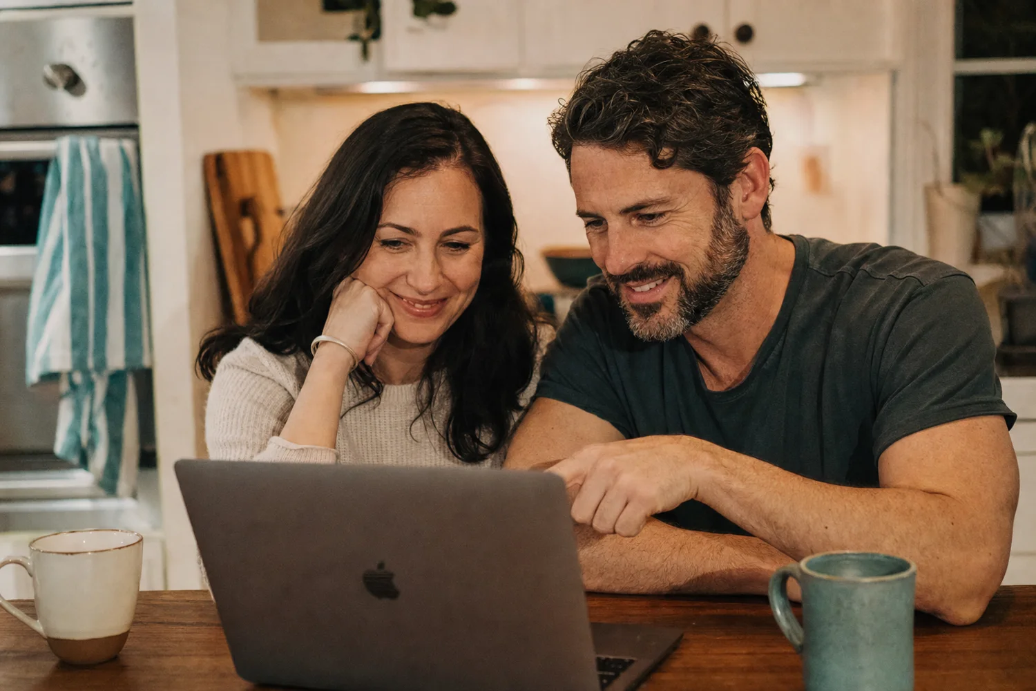 A brother and sister look at a laptop together at a kitchen table.